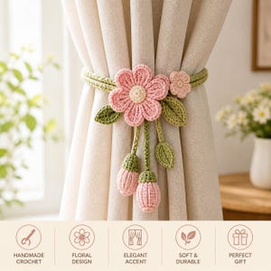 Può includere: Un fermatenda all'uncinetto fatto a mano con un fiore rosa, centro beige, foglie verdi e accenti di boccioli rosa. Il fermatenda è avvolto attorno a una tenda beige, aggiungendo un accento floreale.