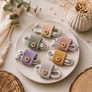 Op de afbeelding: Zes gehaakte kabelorganizers in beige, saliegroen, oudroze, lavendel, mosterdgeel en lichtgrijs. Elke rechthoekige wikkel is voorzien van een kleine, ronde houten knoop met een hartvormige uitsparing, die de witte opgerolde kabels vastzet.