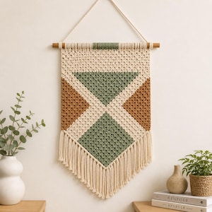 May include: A macrame wall hanging with a geometric design. The piece features a cream-colored background with brown and green diamond shapes. It hangs from a wooden dowel and has a fringe at the bottom.