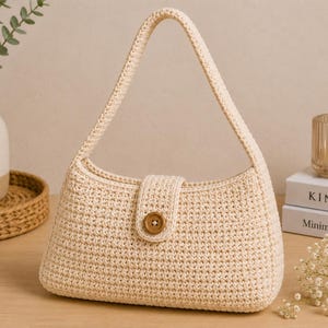 May include: A cream-coloured, crocheted handbag with a curved handle and a flap closure secured by a wooden button. The bag is rectangular with a rounded bottom. The background includes a vase, books, and a candle.