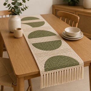 Peut inclure: Chemin de table en crochet beige et vert olive avec un motif géométrique. Le chemin de table présente des demi-cercles vert olive, séparés par des rayures horizontales. Les extrémités ont des franges. Posé sur une table en bois.