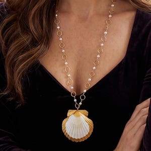 May include: A long gold-tone necklace with a large seashell pendant. The pendant is white with gold trim. The necklace has small pearls and gold circles. The woman is wearing a dark purple velvet top.