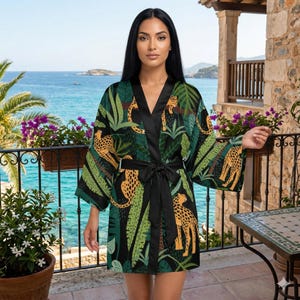 Op de afbeelding: Een kimono-stijl badjas met een levendige jungleprint met gele en zwarte luipaarden tussen groene palmbladeren en gebladerte. Het kledingstuk heeft een effen zwarte, satijnachtige bies langs de opening en een bijpassende zwarte ceintuur.