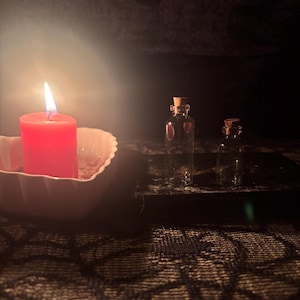 Puede incluir: Una vela de pilar roja se encuentra dentro de un cuenco blanco en forma de corazón junto a dos botellas de vidrio transparente pequeñas con tapones de corcho. La escena está iluminada por la llama cálida de la vela sobre los frascos.