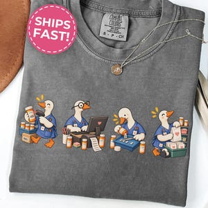 May include: A charcoal gray t-shirt featuring a graphic of four white ducks dressed in blue pharmacy scrubs. The ducks are shown performing various tasks including scanning medication, typing on a computer, counting pills, and bagging prescriptions.