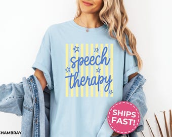 May include: A light blue t-shirt featuring a graphic with vertical yellow stripes and the blue text "speech therapy" written in a cursive font. The design is accented with small blue star outlines scattered around the text.