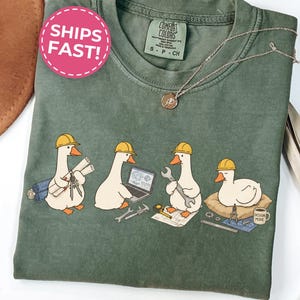 May include: A moss green t-shirt featuring a graphic of four white geese wearing yellow construction hard hats. The geese are depicted as engineers with blueprints, a laptop, a wrench, and a coffee mug labeled Design Mode on a pillow.