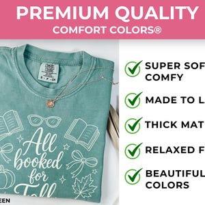 May include: A light green Comfort Colors t-shirt featuring white line art of books, glasses, bows, a pumpkin, and a maple leaf with the text All booked for Fall. The shirt includes a Comfort Colors brand tag and a delicate gold-toned necklace.