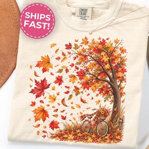 May include: An ivory t-shirt featuring a graphic print of a tree with vibrant red, orange, and yellow autumn leaves blowing in the wind. A bicycle with a basket of sunflowers is parked at the base of the tree, surrounded by fallen leaves.