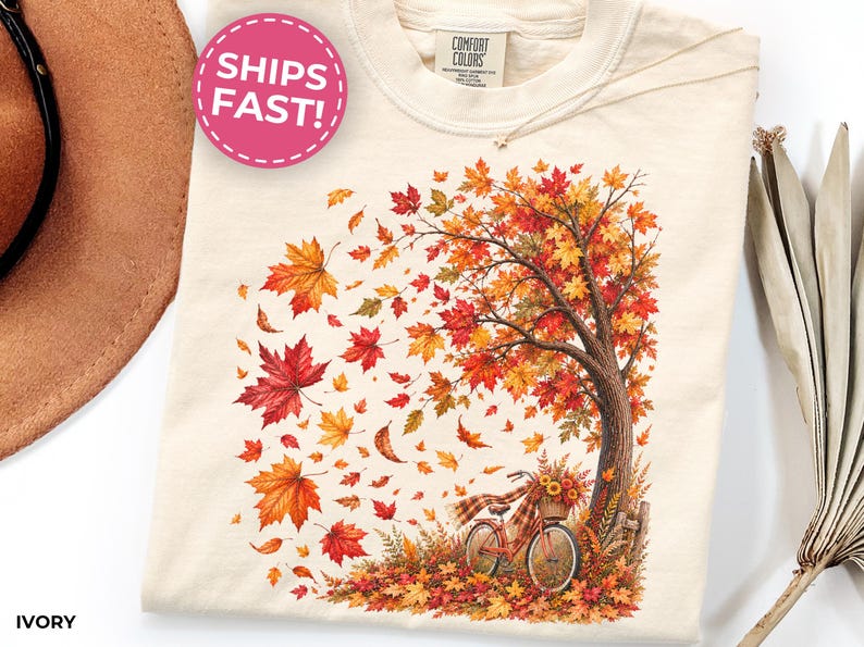 May include: An ivory t-shirt featuring a graphic print of a tree with vibrant red, orange, and yellow autumn leaves blowing in the wind. A bicycle with a basket of sunflowers is parked at the base of the tree, surrounded by fallen leaves.