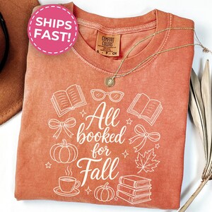 May include: A rust-colored short-sleeve t-shirt featuring a white graphic print with the text All booked for Fall. The design includes illustrations of open books, a stack of books, pumpkins, a maple leaf, eyeglasses, and bows.