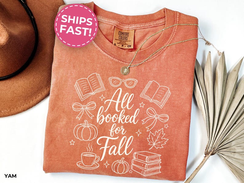 May include: A rust-colored short-sleeve t-shirt featuring a white graphic print with the text All booked for Fall. The design includes illustrations of open books, a stack of books, pumpkins, a maple leaf, eyeglasses, and bows.