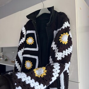 Puede incluir: Una chaqueta negra con paneles de cuadrados de ganchillo en marrón oscuro, blanco y amarillo. La chaqueta tiene un cuello y mangas de color negro sólido, con el patrón de ganchillo integrado en la parte delantera y en las secciones de las mangas.