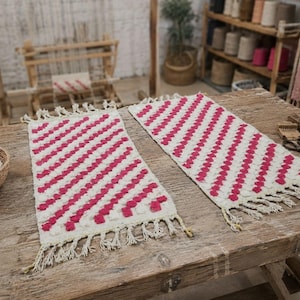 May include: Two rectangular woven table runners featuring a cream-colored base with diagonal stripes of raised, textured hot pink tufted dots. Each runner is finished with short, knotted fringe along the two shorter ends of the fabric.
