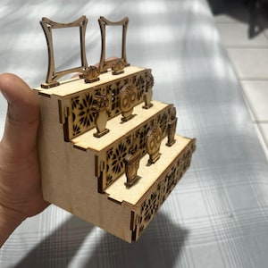 Puede incluir: Un expositor de madera de tres niveles con patrones florales cortados con láser en los laterales. El nivel superior sostiene dos marcos cuadrados, mientras que los niveles inferiores presentan adornos de madera circulares y en forma de jarrón.