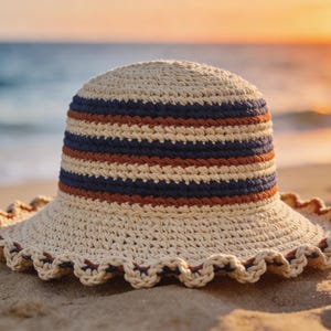 May include: A cream-colored crochet sun hat with a wide, scalloped brim. The hat features horizontal stripes in navy blue and rust-orange around the crown. The hat is resting on a sandy beach with the ocean in the background.