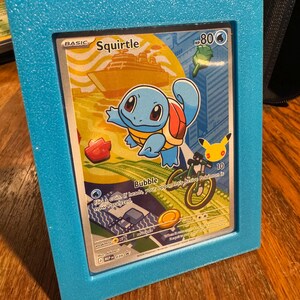 May include: A framed Squirtle Pokémon card. The card features a blue Squirtle character with a red shell, set against a yellow and orange background. The frame is a textured, light blue. The card has the text "Squirtle" and "Bubble".