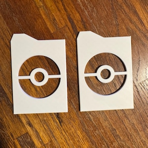 May include: Two white, rectangular card holders with a circular design cut out, resembling a stylized ball. The design features a central circle bisected by a horizontal line. The card holders are placed on a wooden surface.