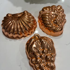 May include: Three copper-colored metal baking molds with a shiny, reflective finish. One mold is shaped like a seashell with radiating ridges, while the other two molds feature detailed, embossed designs of clusters of grapes and leaves.