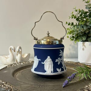 May include: A navy blue ceramic jar with a silver handle and lid. The jar features white bas-relief figures and foliage. The jar is displayed on a silver-colored tray, with white swan figurines and greenery in the background.