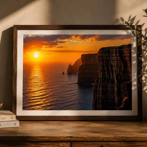 Könnte beinhalten: Gerahmter Landschaftsdruck mit Sonnenuntergang über dem Meer. Die Sonne spiegelt sich im Wasser. Auf der rechten Seite sind Klippen zu sehen. Der Rahmen ist dunkelbraun.