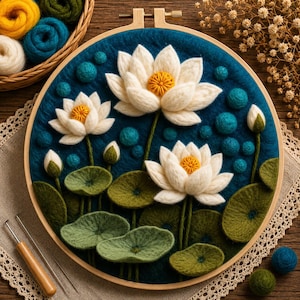 Puede incluir: Un bastidor de bordado artesanal con flores de loto blancas de fieltro con centros amarillos, hojas de nenúfar verdes y detalles turquesa sobre un fondo azul oscuro. Diseño floral y artesanal.