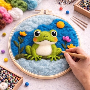 Puede incluir: Bordado de rana de fieltro hecho a mano en un aro de madera. La rana es verde y blanca con grandes ojos negros y está sentada sobre un tronco de fieltro. El fondo presenta un estanque azul, un sol amarillo y flores de colores. Suministros de artesanía dispersos.