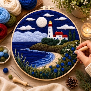 Puede incluir: Una pieza de arte textil hecha a mano que representa un paisaje costero con un faro, el mar y una playa. La obra de arte está enmarcada en un aro de madera y presenta colores azules, blancos, verdes y marrones. También se ven suministros de artesanía, incluyendo lana y agujas.