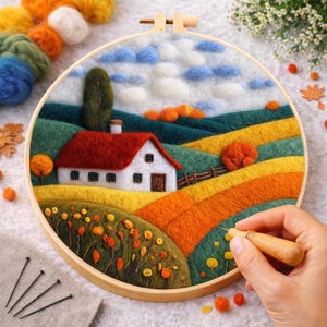 May include: A felt landscape scene in a wooden embroidery hoop. The artwork shows a white house with a red roof, rolling hills in green, yellow, and orange, and a blue sky with white clouds. The scene evokes autumn. The hoop measures approximately 20 cm in diameter.