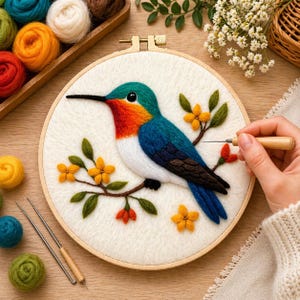 Puede incluir: Un colibrí colorido, realizado con la técnica del fieltro con aguja, dentro de un bastidor de bordado de madera. El ave presenta detalles en azul, verde, naranja y blanco, posado en una rama con flores amarillas y hojas verdes.