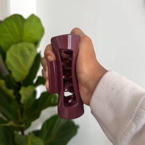 May include: A burgundy hair claw clip, held in a hand, with a curved design and interlocking teeth. The clip is made of a solid, smooth material and is designed to hold hair in place. The background includes a green plant and a white wall.
