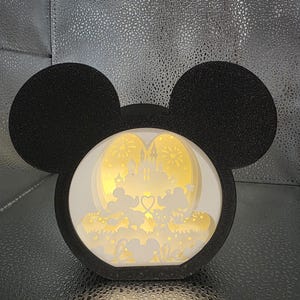 May include: A black Mickey Mouse-shaped light box with a layered design. Inside, silhouettes of Mickey and Minnie Mouse are seen in front of a castle, with the word "love" visible. The light box is a decorative item.