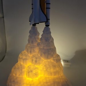 May include: A white and orange rocket ship lamp with a glowing, textured base. The rocket has black accents and is positioned as if launching. The base emits a warm, yellow light, creating a space-themed decorative piece.