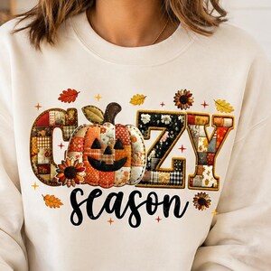 以下が含まれることがあります： 黒い筆記体で「cozy season」という文字がプリントされた、クリーム色のクルーネックのスウェットシャツ。「cozy」の文字は、中央に配置されたカボチャと、オレンジ、茶色、黄色の色調のパッチワーク模様でデザインされています。