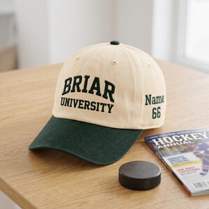 May include: A cream-colored baseball cap with a dark green brim. The front features dark green embroidered text that reads BRIAR UNIVERSITY. The side of the cap includes dark green embroidered text that reads Name and the number 66.