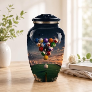 May include: A dark blue cremation urn with a glossy finish, featuring a surreal design of a pool table scene. The urn showcases a triangle of billiard balls floating against a sunset sky, with a cue stick and a white cue ball.