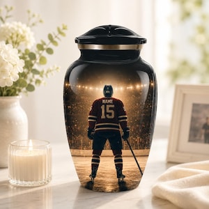 Può includere: Urna funeraria nera con una fascia dorata e un design a giocatore di hockey. L'urna presenta un giocatore di hockey in una maglia bordeaux e oro con il numero 15 e il nome "NAME" sulla schiena, in piedi sul ghiaccio in uno stadio.