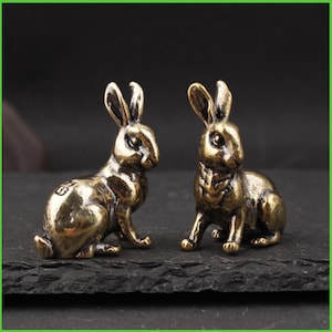 May include: Two bronze-colored rabbit figurines. The rabbits are in a seated position with their ears perked up. The figurines have detailed features, including eyes, noses, and paws. The rabbits are displayed on a dark surface.