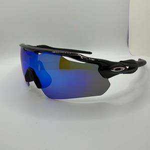 May include: Black performance sunglasses featuring a single-lens shield design with a reflective blue and purple gradient finish. The frame includes a black nose bridge and temples with the Oakley logo visible on the side hinge area.