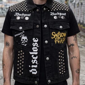 Puede incluir: Un chaleco de mezclilla negro con tachuelas metálicas plateadas en los hombros y paneles frontales. La prenda muestra texto blanco que dice Blackgoat y disclose, un gráfico de calavera blanca y texto amarillo Shadow of Fear en los bolsillos.