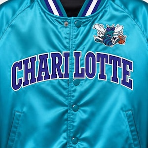 May include: A teal satin jacket with the word "CHARLOTTE" in white and blue lettering across the chest. The jacket has a logo of a cartoon bee with a basketball. The jacket has white and blue striped trim on the collar, cuffs, and hem.