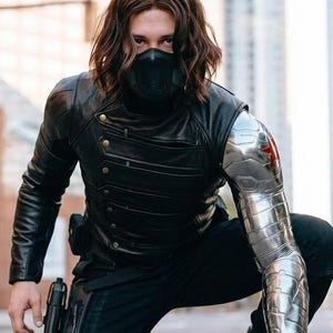 Op de afbeelding: Een persoon in een zwart leren jack, broek en masker, met een metalen arm en een pistoolholster. De persoon knielt op een stadsstraat. De metalen arm heeft een rode ster.