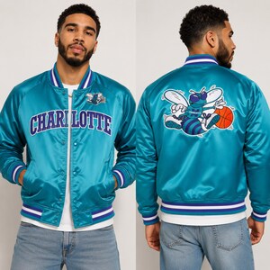 May include: Teal satin bomber jacket with the word "CHARLOTTE" across the chest. The jacket features a white and purple striped collar, cuffs, and hem. The back of the jacket displays a cartoon bee holding a basketball.