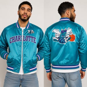 May include: Teal satin bomber jacket with the word "CHARLOTTE" across the chest. The jacket features a white and purple striped collar, cuffs, and hem. The back of the jacket displays a cartoon bee holding a basketball.