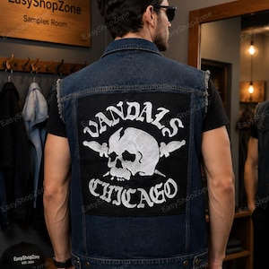 Puede incluir: Un chaleco de mezclilla azul oscuro con un gran parche rectangular negro en la espalda. El parche muestra un gr&aacute;fico blanco de una calavera con dos cuchillos cruzados y el texto VANDALS CHICAGO en una fuente llamativa. El chaleco tiene sisas deshilachadas.