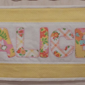 May include: A yellow and white fabric banner with the name "ALICE" spelled out in colorful fabric letters.