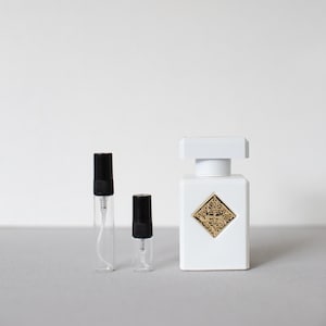 May include: A white perfume bottle with a gold emblem and two clear sample vials with black caps. The perfume bottle has a square shape with a white cap. The vials are different sizes.