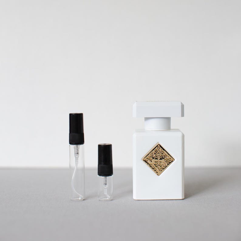 May include: A white perfume bottle with a gold emblem and two clear sample vials with black caps. The perfume bottle has a square shape with a white cap. The vials are different sizes.