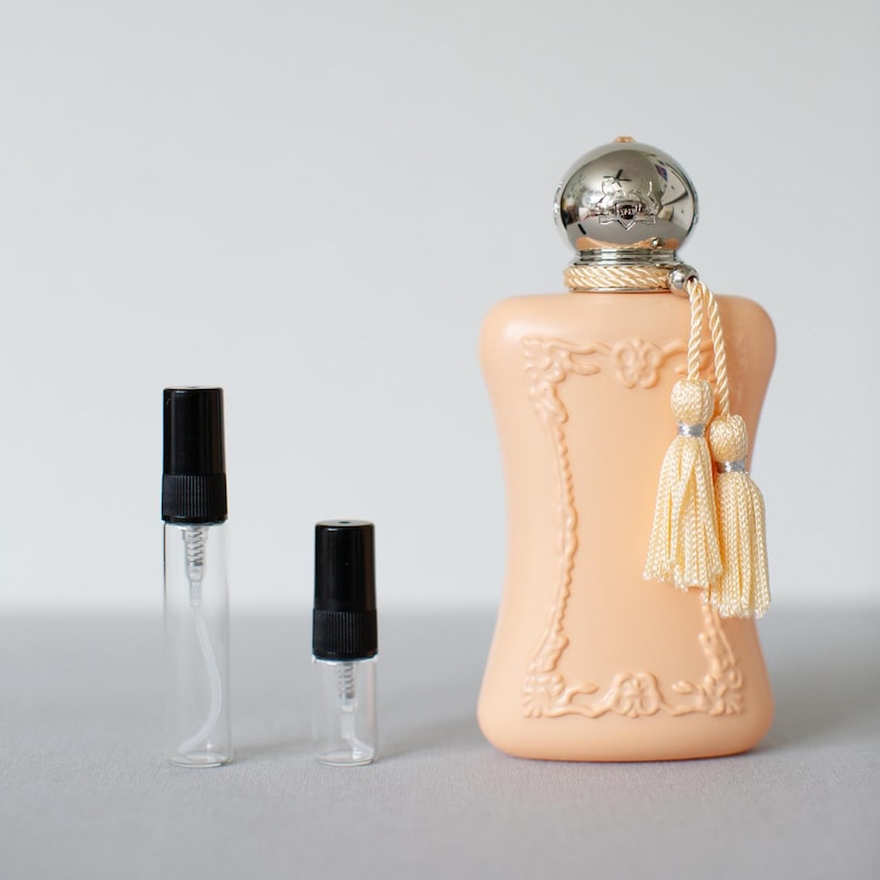 May include: A peach-coloured perfume bottle with a silver cap and two cream-coloured tassels, accompanied by two small, clear glass sample vials with black spray caps. The bottle features an ornate, raised frame design on its front surface.
