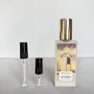 May include: A rectangular glass perfume bottle with a gold cap and a label featuring a lighthouse illustration and the text Memo Paris Cap Camarat Eau de Parfum, alongside two smaller empty glass sample vials with black spray caps.